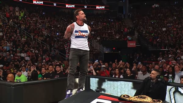 A wrestler stands confidently on an announce table in a packed arena, preparing to disrobe. The audience is engaged, capturing the moment with their phones.