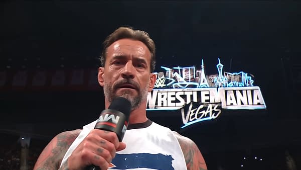 CM Punk stands on stage speaking into a microphone, expressing his views while a large WrestleMania logo is illuminated behind him.