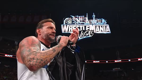 A man with tattoos, speaking passionately while holding a black jacket, stands in front of a vibrant WrestleMania sign during a WWE Raw event.