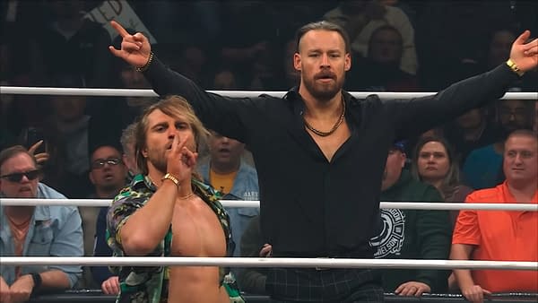 Clark Connors and David Finlay of The Dogs stand confidently in the wrestling ring, with Connors making a shushing gesture and Finlay raising his arms triumphantly. The audience is visible in the background, showing a range of reactions.