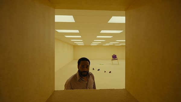 A man peers through a narrow opening into a stark, yellow-lit backrooms-like empty room with fluorescent ceiling panels and a lone purple chair in the distance.