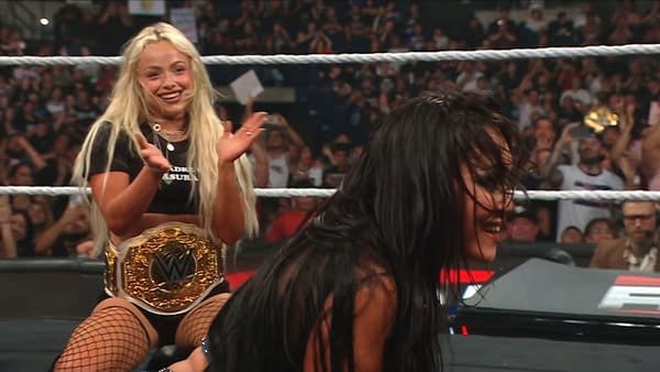Liv Morgan smiles and applauds Roxanne Perez in the WWE Raw ring, with a championship belt around her waist and the crowd in the background.
