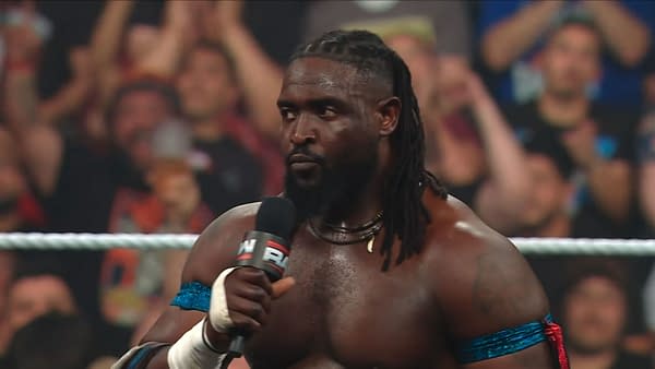 Oba Femi stands in the WWE Raw ring holding a microphone, staring intensely toward an unseen challenger. The crowd blurs in the background.