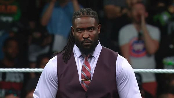 A man with long braided hair wearing a dark vest and light purple shirt stands confidently in a wrestling ring, appearing serious against a cheering crowd in the background. He is dressed for a formal occasion, enhancing his commanding presence.