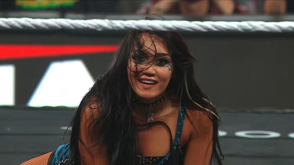 Roxanne Perez crouches in the WWE Raw ring, smiling with her hair falling across her face. She wears a blue ring outfit and dark eye makeup.