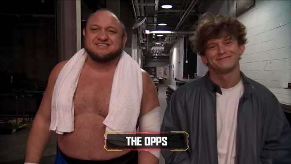 Samoa Joe and Hook of The Opps smile while walking backstage on AEW Collision. They are in a narrow hallway, with Samoa Joe wearing a towel around his neck.