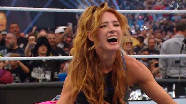 A woman's face filled with joy and excitement, her long, wavy red hair framing her smile. She is surrounded by an enthusiastic crowd, celebrating a victory in a wrestling event.