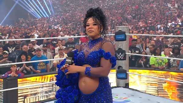 A woman stands in a wrestling ring wearing a stunning blue outfit adorned with flowers, holding a microphone. The audience is visible in the background, showcasing a vibrant WrestleMania atmosphere.