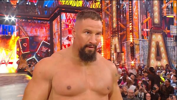 A muscular man with a beard stands in a wrestling ring, focused on the action, surrounded by an excited crowd and colorful lighting from the event's backdrop.