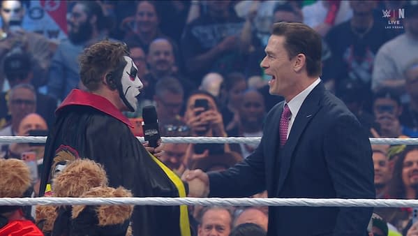 A performer in face paint and a cape with colorful details shakes hands with a man in a suit, surrounded by a cheering crowd at a wrestling event. Spotlight on the performers highlights the energetic atmosphere.