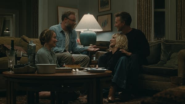 A still from 'The Invite' showing four friends sitting in a cozy living room, laughing and engaging in conversation. The setting features a warm atmosphere with a table holding drinks and snacks, and a lamp illuminating the scene.