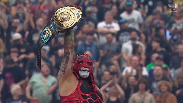 A masked wrestler in a vibrant red outfit triumphantly holds up a championship belt above their head in front of a cheering crowd. The background shows enthusiastic fans celebrating the moment.