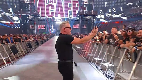 A man in sunglasses and a black shirt gestures to the crowd while walking down a wrestling arena walkway, with a large screen displaying his name in the background and a crowd of fans visible.