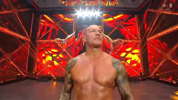 A muscular man stands confidently in a wrestling arena, illuminated by dramatic red lighting and spotlights. His tattooed arms are visible, and he faces the crowd with a determined expression.