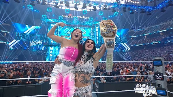 Two women celebrate in a wrestling ring at WrestleMania, with one holding a championship belt high. The background features bright lights and a cheering crowd.