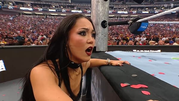 A shocked woman with dark hair and dramatic makeup watches with concern at a wrestling event, surrounded by a cheering crowd in the background. Her expression conveys surprise and suspense as the action unfolds.