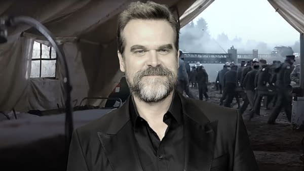 David Harbour smiles while dressed in a black suit at the Critics Choice Awards 2025, with a blurred background featuring a scene possibly related to a film set.