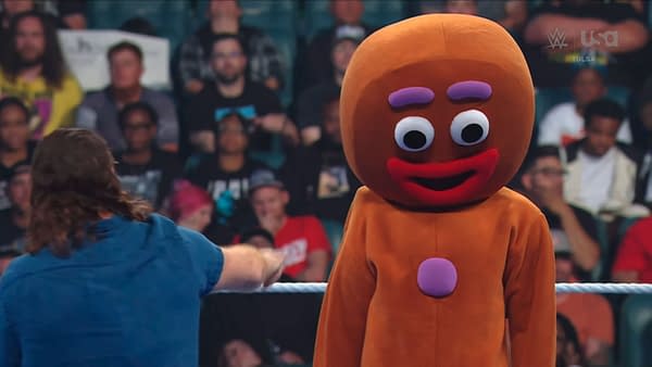 Sami Zayn points toward a person in a giant gingerbread man costume standing helplessly in the WWE ring. The crowd watches in the background.