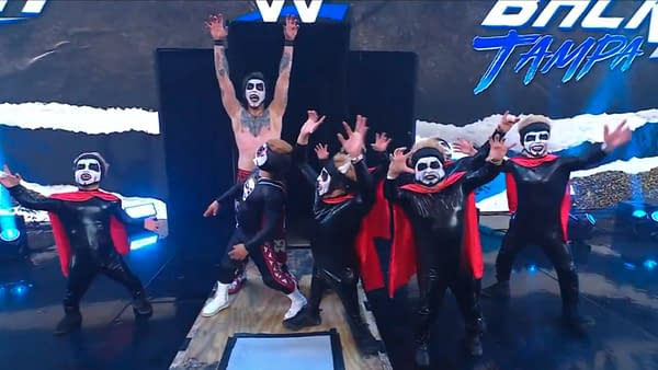 Danhausen stands on the WWE Backlash stage with several masked dancers or performers in black-and-red costumes, raising his arms in a celebratory pose.