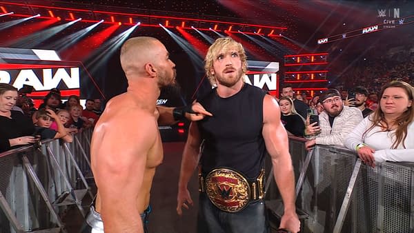 Logan Paul stands in the WWE Raw arena wearing a championship belt while another wrestler confronts him at ringside. Fans watch from behind the barricade.