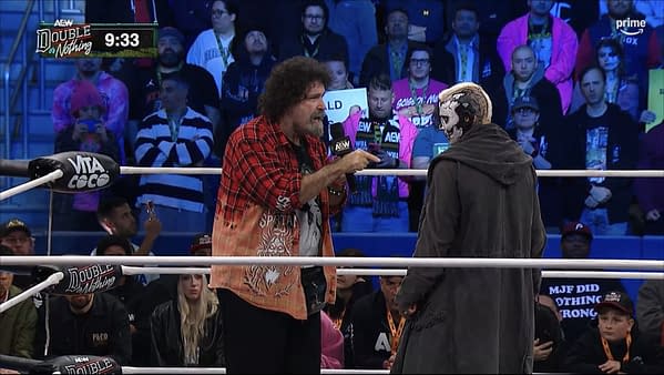 Mick Foley, in a red plaid shirt, points and speaks to Darby Allin during the AEW Double or Nothing Buy-In pre-show inside the wrestling ring. The crowd watches from the stands behind them.