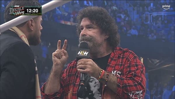 Mick Foley speaks into an AEW microphone while facing MJF and holding up two fingers at AEW Double or Nothing. The Buy-In pre-show crowd blurs in the background.