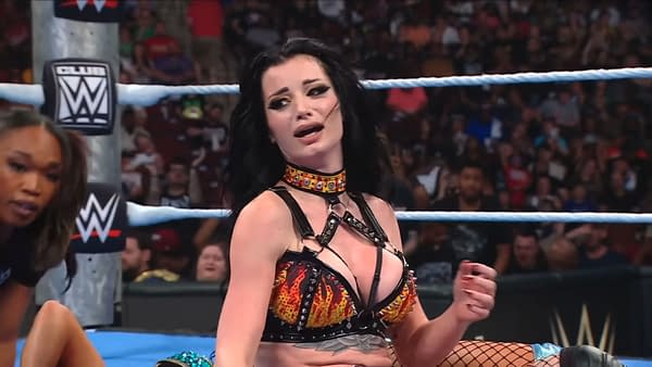 Paige stands in the WWE ring celebrating a hard-fought SmackDown victory. An opponent is down beside her as the crowd watches in the background.