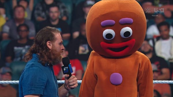 Sami Zayn speaks into a microphone beside a person in a giant gingerbread man costume on WWE SmackDown.