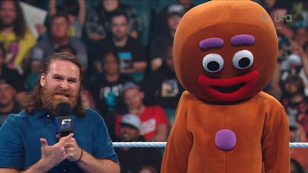 Sami Zayn stands in the WWE SmackDown ring beside a giant gingerbread man costume, speaking into a microphone. The crowd blurs in the background.