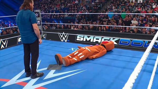 A WWE wrestler stands in the ring facing a person in a Gingerbread Man costume lying on the mat during a SmackDown segment. The crowd watches as the scene builds tension.