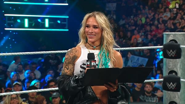 Sol Ruca smiles in the WWE ring while holding a contract during a live WWE Raw segment. The crowd and ring ropes are visible behind her.