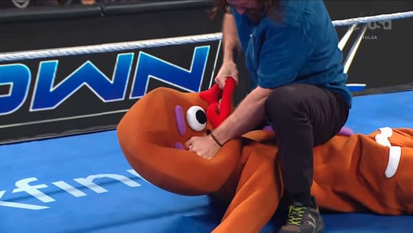 Sami Zayn kneels over an empty Gingerbread Man costume and grapples with it on the WWE SmackDown ring mat.