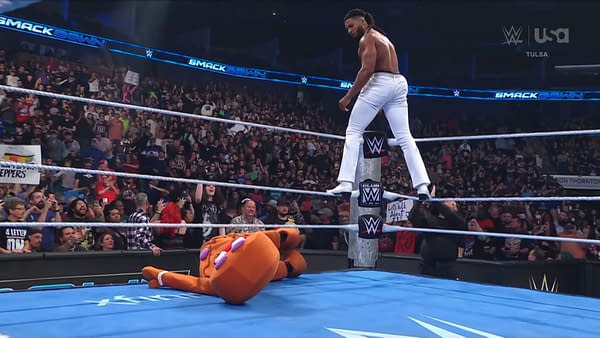 Trick Williams stands on the turnbuckle in a WWE SmackDown ring, looking down at a damaged gingerbread-man figure lying on the mat after a Sami Zayn attack. The crowd fills the arena behind him.