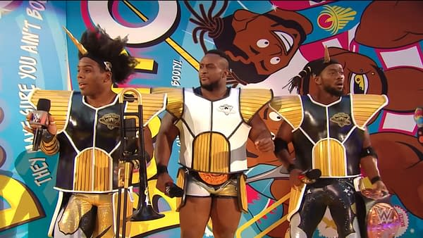 Xavier Woods, Big E, and Kofi Kingston of The New Day stand in colorful wrestling gear with microphones in front of a bright cartoon-style backdrop.