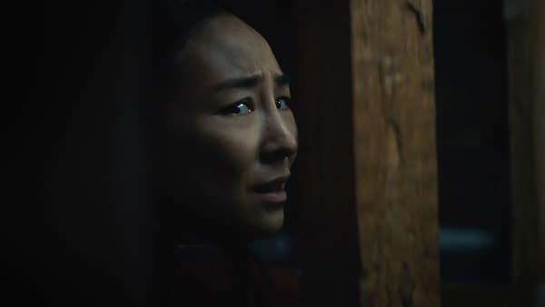 Greta Lee as Ann in Hideaway, shown in a tense close-up peering anxiously from the shadows beside a wooden beam.