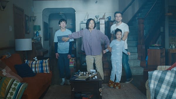Four people stand tensely in a dim family living room in The Last House, with a staircase, sofa, and chessboard in the foreground.
