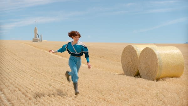 A young woman runs through a golden wheat field beside large round hay bales under a bright blue sky.