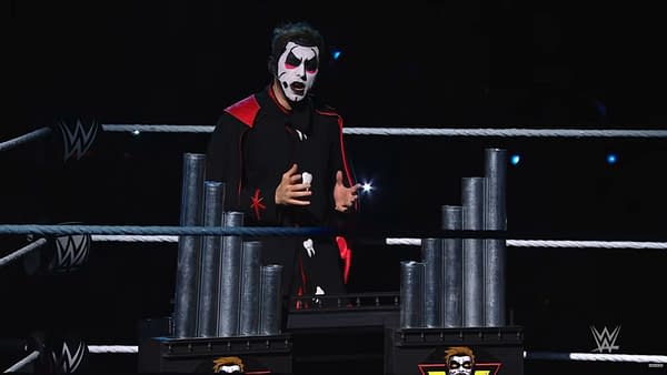 Danhausen stands in a WWE Raw ring, wearing dramatic face paint and a black-and-red outfit while performing on a dark stage. He is framed by ring ropes and metallic set pieces.