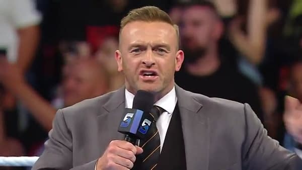 A close-up shot of WWE SmackDown General Manager Nick Aldis, branded microphone in hand, addressing the crowd from within the wrestling ring at the start of the show. A out-of-focus crowd is visible in the background.
