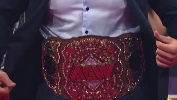 A close-up of the AEW Championship title belt around the waist of Kenny Omega, who holds his suit jacket open to expose the gold belt.