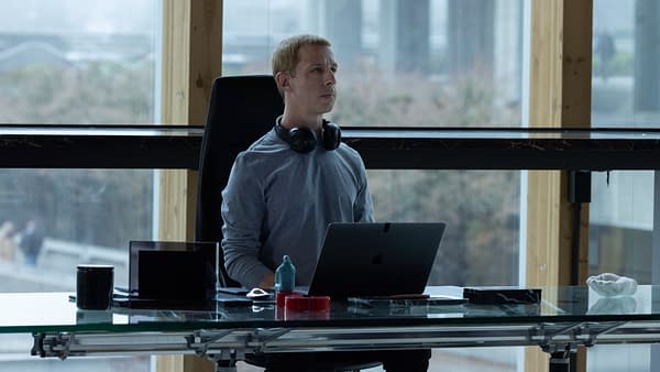 Man wearing headphones sits at a laptop in a modern glass-walled office, looking contemplative.