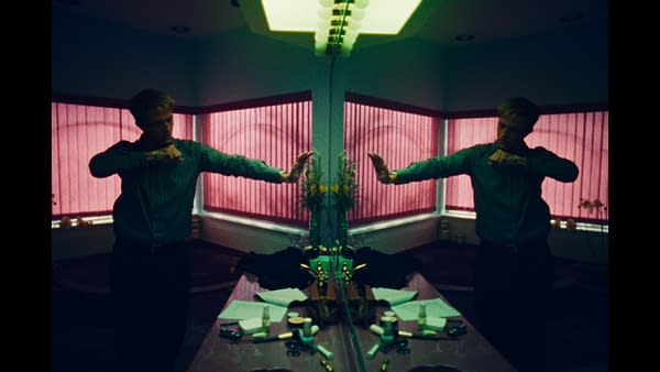 A mirrored, dimly lit bathroom scene shows a person standing with arms raised in front of a vanity, with pink blinds and scattered toiletries.