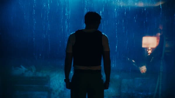 A silhouetted man stands in heavy rain, lit by blue light and a glowing streetlamp, creating a moody primetime scene.