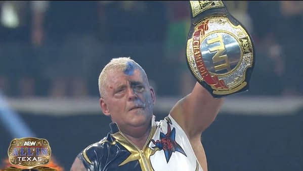Dustin Rhodes, wearing a star-themed outfit and with blue face paint, celebrates triumphantly as he raises the TNT Championship belt high above his head after winning at AEW All In: Texas.