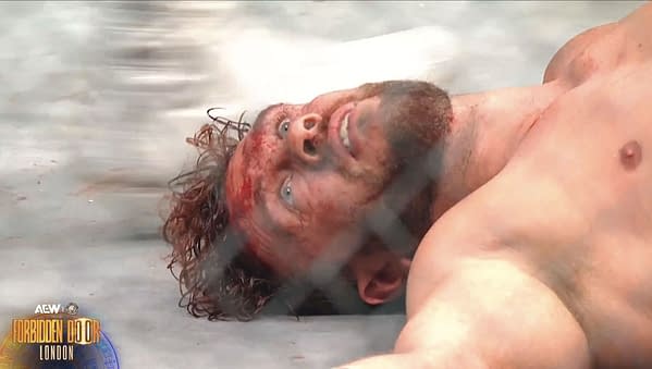 Will Ospreay, a wrestler, lies on the mat in a state of distress after a brutal Lights Out Steel Cage Match, his face covered in blood and showing visible fatigue. The scene is set against the backdrop of a steel cage.
