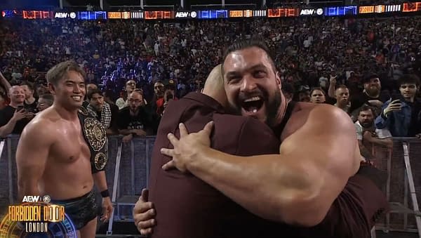 Wardlow embraces Don Callis with joy, while Kazuchika Okada observes in the background at the Forbidden Door event. The audience can be seen enthusiastically reacting to the moment.