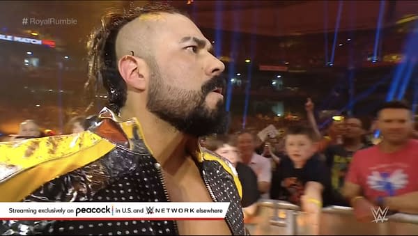 A wrestler with a distinctive mohawk hairstyle and a colorful jacket enters the Royal Rumble arena, where fans are cheering and reaching out to him. The energy of the crowd is palpable, with bright lights illuminating the scene.
