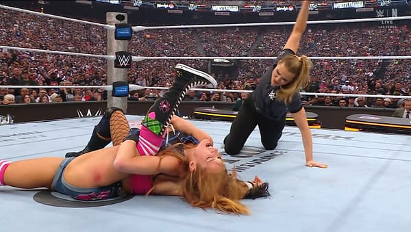 In a wrestling ring, Becky Lynch is performing a hold on her opponent, showcasing determination and skill. The audience in the background is visibly engaged, cheering on the action.
