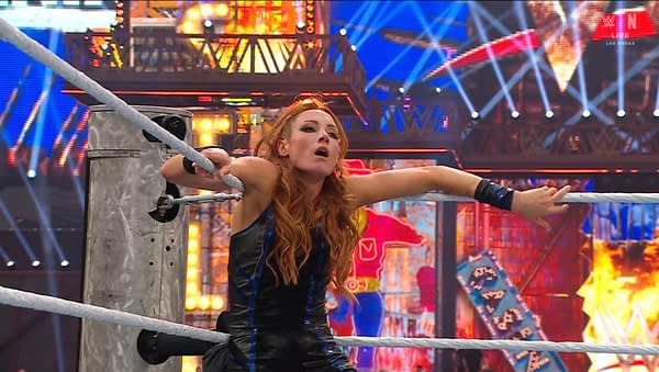A wrestler with long red hair and a black outfit leans against the ropes in a wrestling ring, showing emotion after a match, with bright lights and colorful arena decorations in the background.