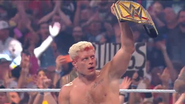 Wrestler Cody Rhodes, with a bloody face and a winning expression, raises the WWE Championship belt high above his head while a cheering crowd surrounds him.